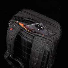 A smartphone partially peeks from the top pocket of a black GORUCK GR2 backpack against a stark black background, showcasing its durable design inspired by Special Forces medical packs.