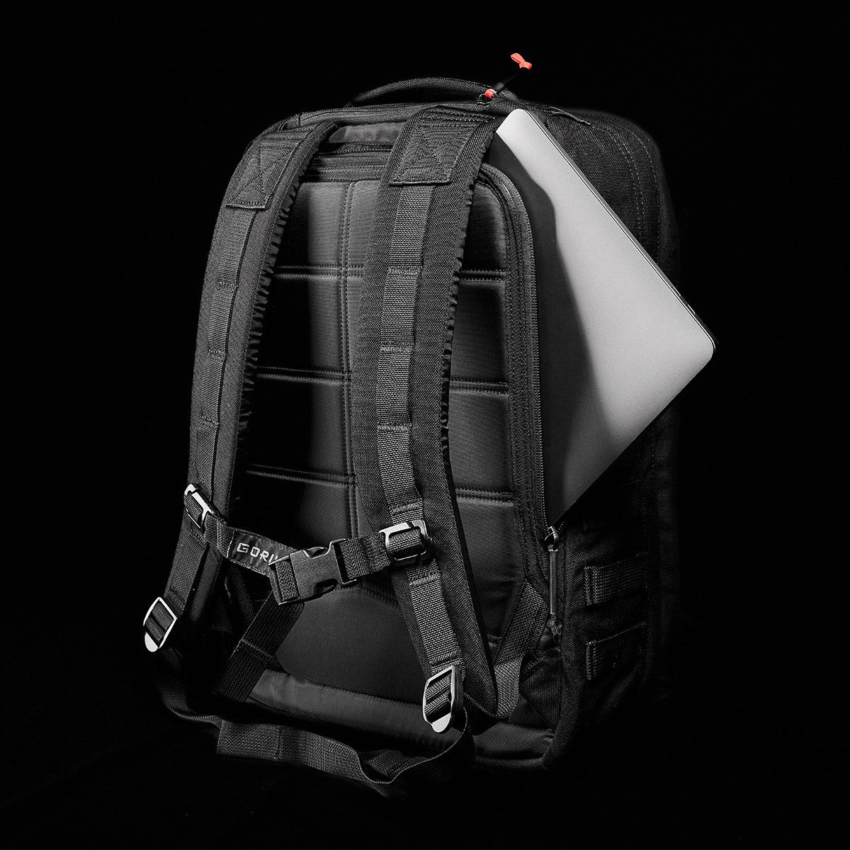 A GORUCK GR2 black backpack, designed to resemble Special Forces medical packs, reveals a laptop emerging from the back compartment against a dark background.