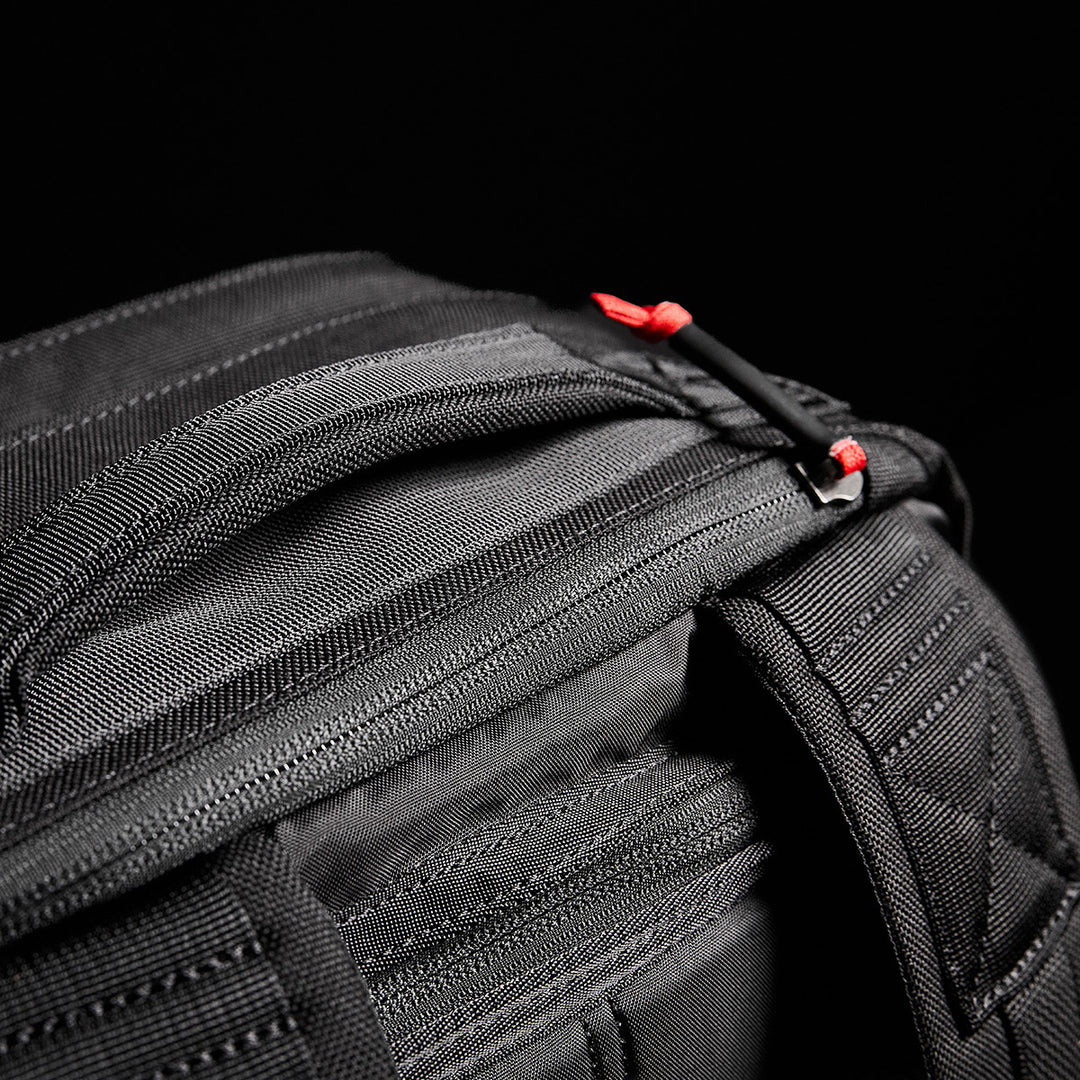Close-up of a black GR2 fabric bag by GORUCK, reminiscent of Special Forces medical packs, featuring red zipper pulls on a dark background.