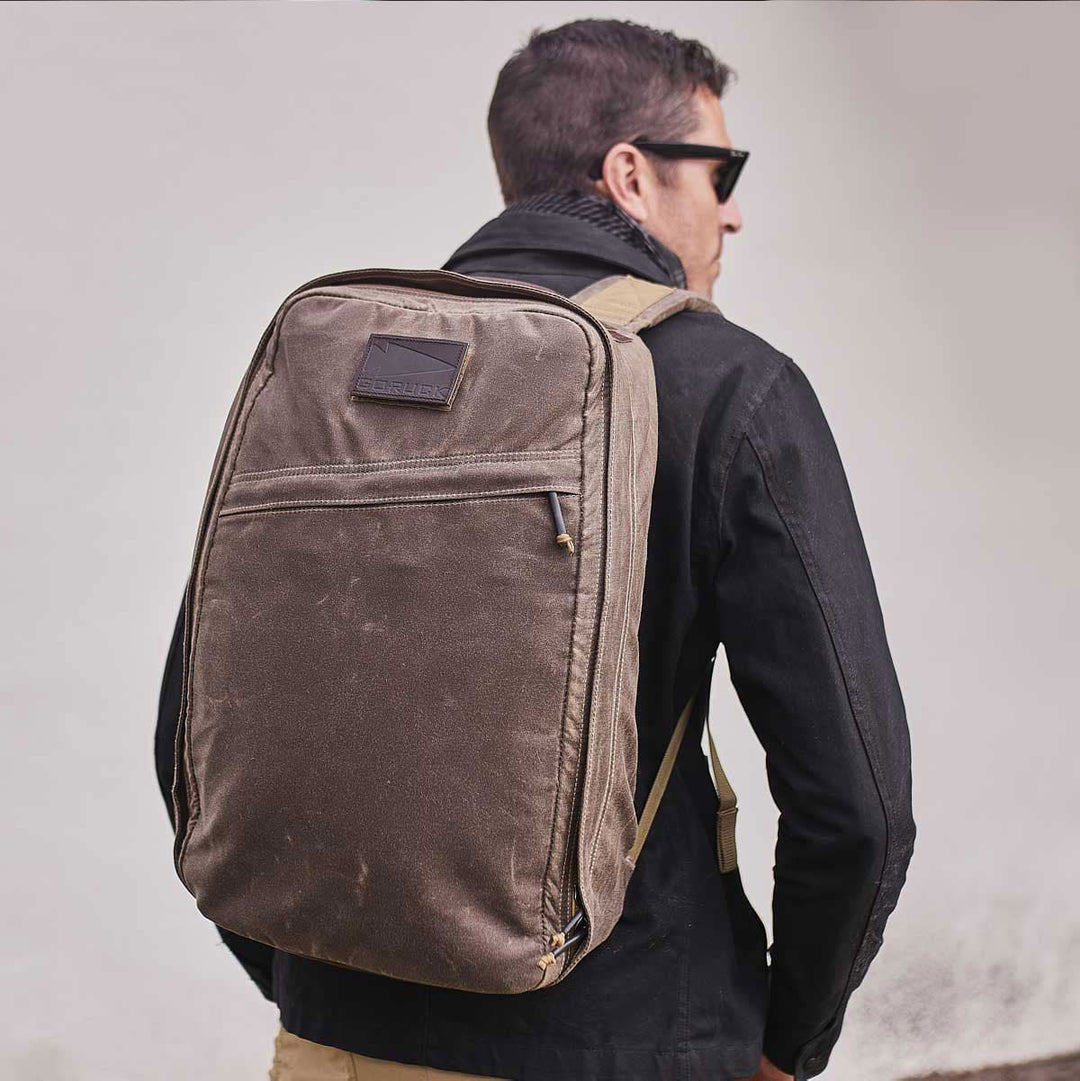 A man wearing sunglasses and a black jacket strides confidently, carrying the GR1 USA - Heritage rucksack from GORUCK, crafted from waxed duck canvas.