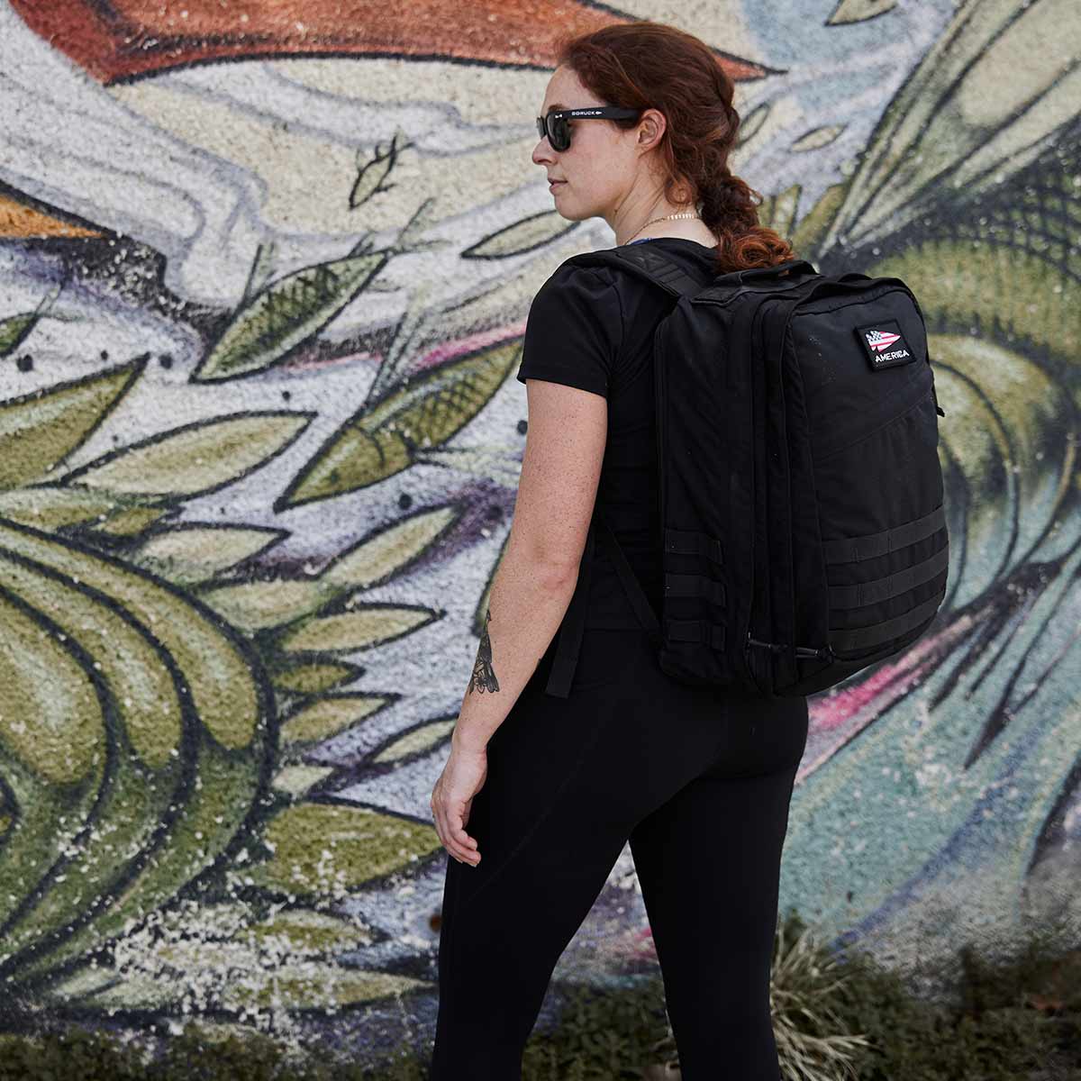 A woman with a black GR2 backpack from GORUCK stands in front of a colorful mural, wearing sunglasses and black clothing. The backpack, renowned for its Scars Lifetime Guarantee, perfectly complements her style.