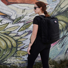A woman with a black GR2 backpack from GORUCK stands in front of a colorful mural, wearing sunglasses and black clothing. The backpack, renowned for its Scars Lifetime Guarantee, perfectly complements her style.