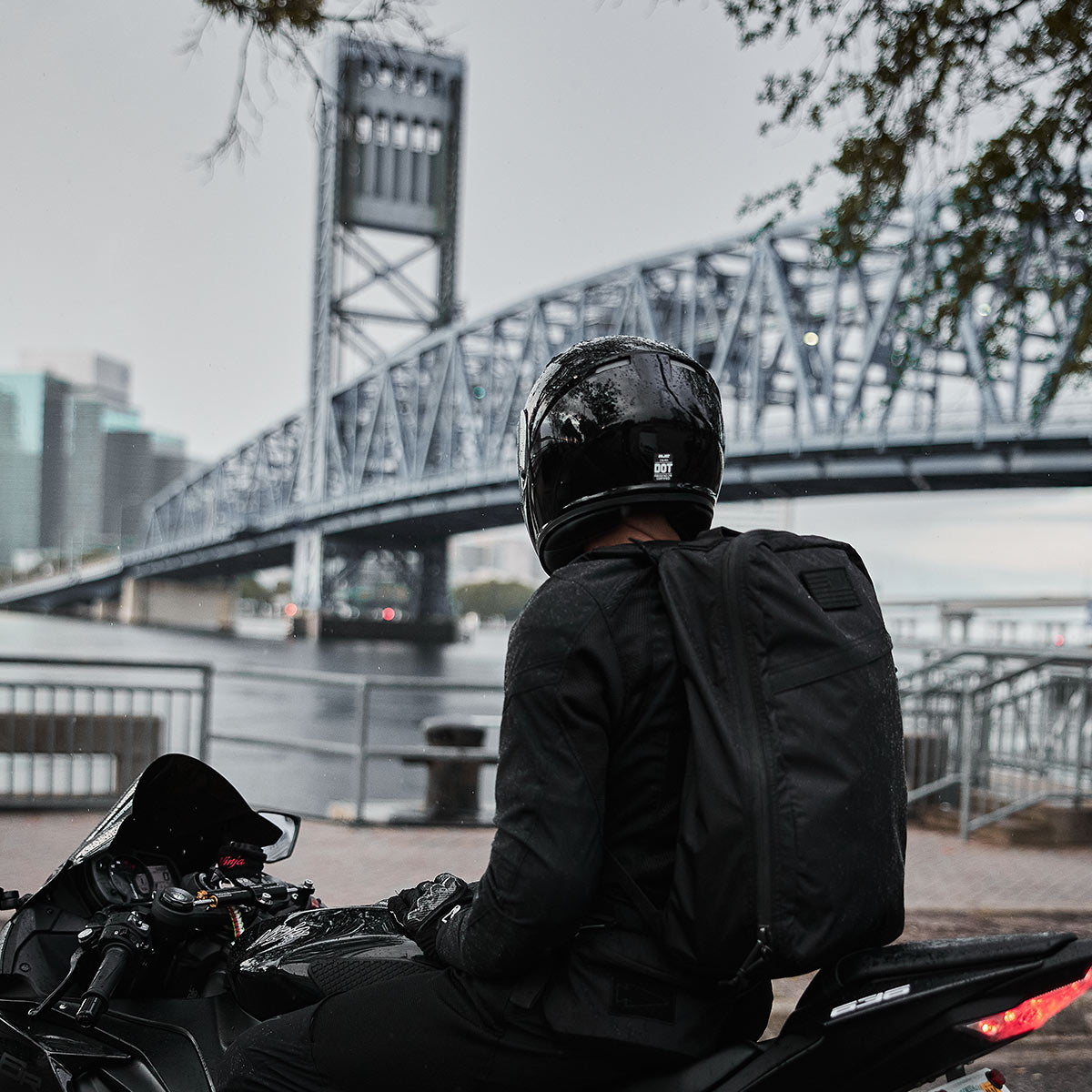 A person in black gear and helmet sits on a motorcycle near a river with a large steel bridge in the background.