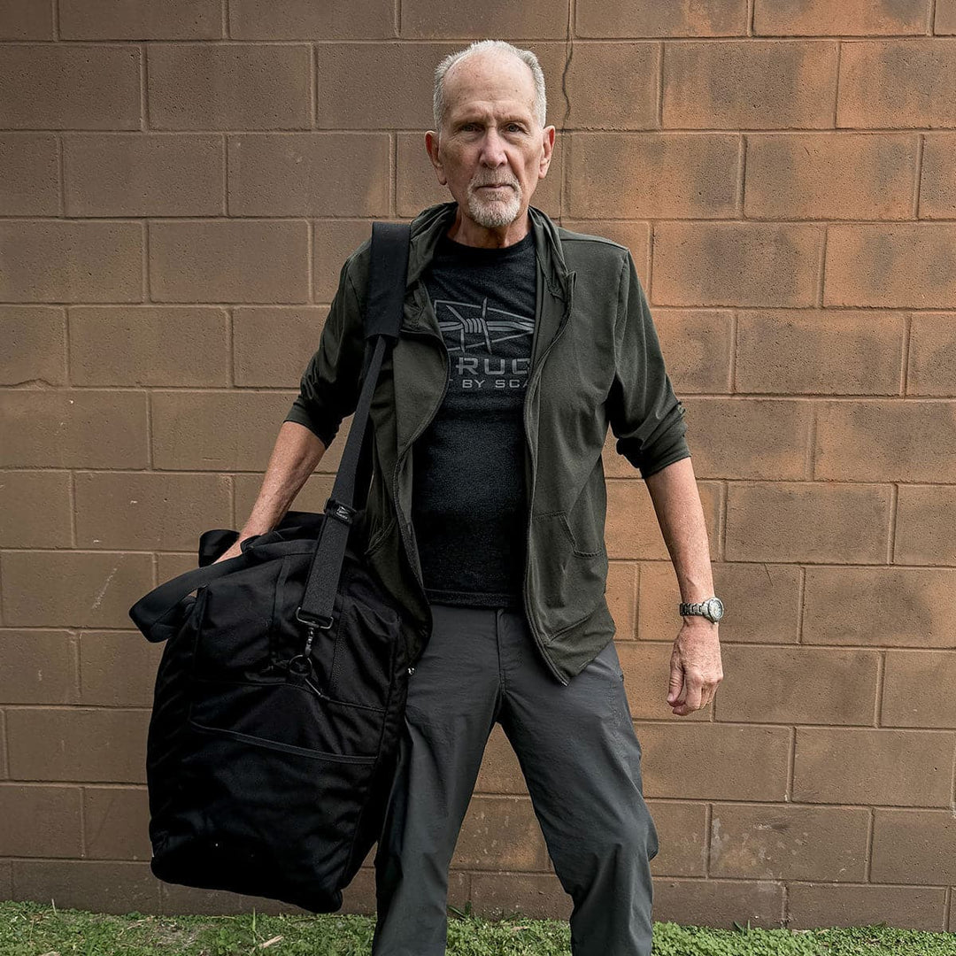 A man stands against a brick wall, holding a large GORUCK Kit Bag - 84L (Includes Shoulder Strap), dressed in rugged outdoor clothing.
