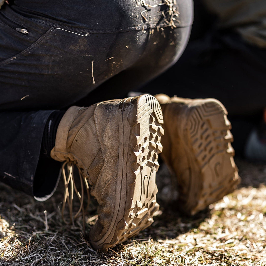 A close-up shot captures a person kneeling on grass, sporting black pants and GORUCK's MACV-2 - Mid Top - Coyote hiking boots with a triple compound outsole. The rugged soles display visible dirt as sunlight casts shadows and highlights the details of the boots and ground.