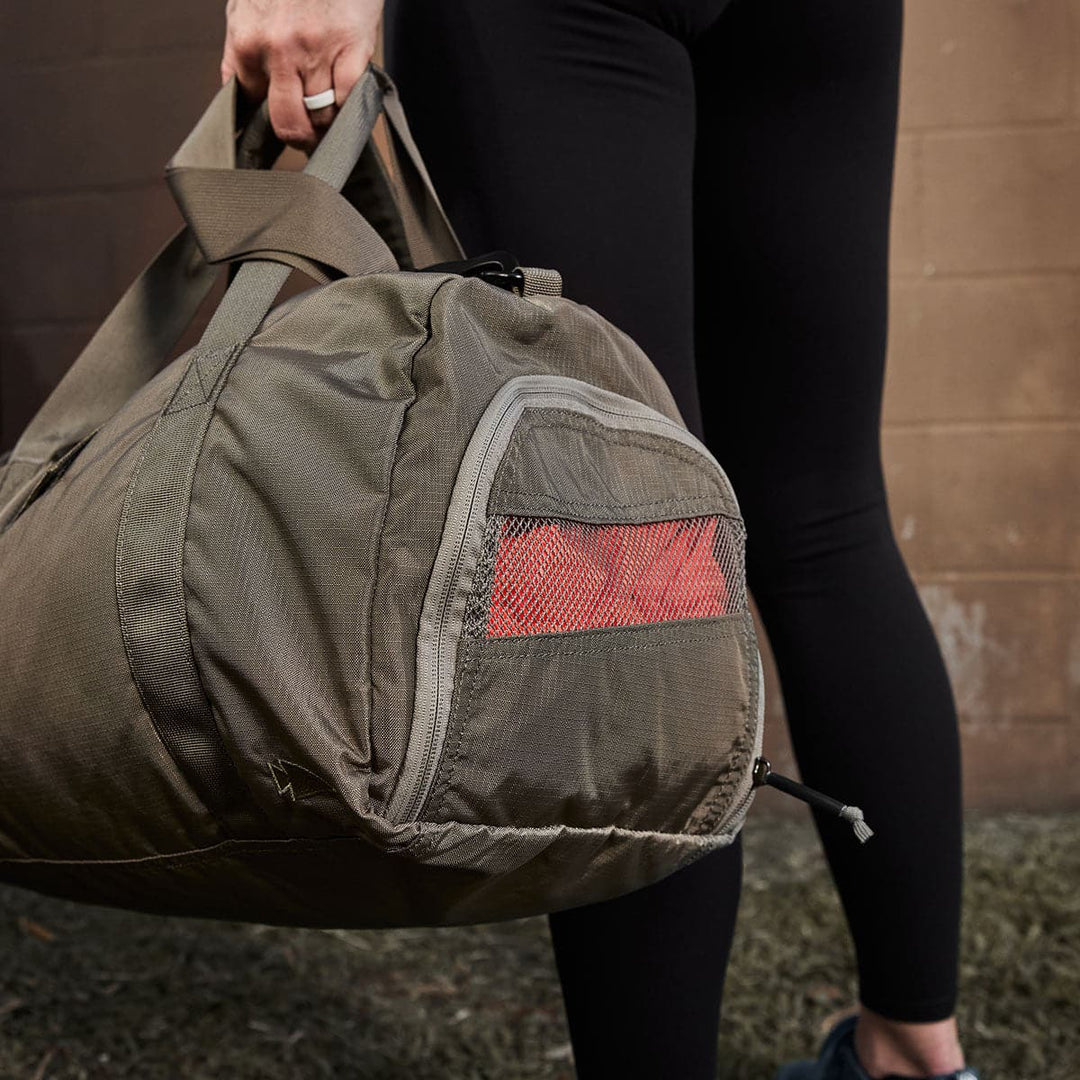 A person stands on the grass, dressed in black leggings and holding a GORUCK Gym Bag - Ripstop ROBIC®, which features a handy shoe compartment.