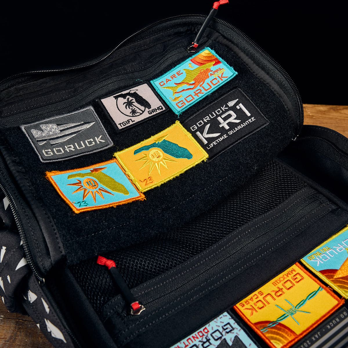A black GORUCK KR1 - Spearhead Splatter kids' rucksack is displayed open on a wooden surface, featuring various colorful patches inside. The patches include abstract designs, logos, and text such as 