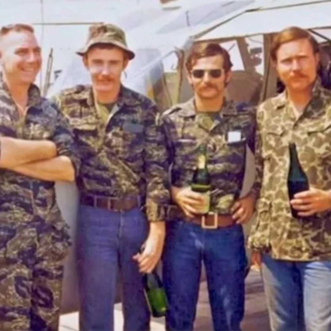 Four men in camouflage uniforms stand by a helicopter, smiling and holding bottles in their hands.