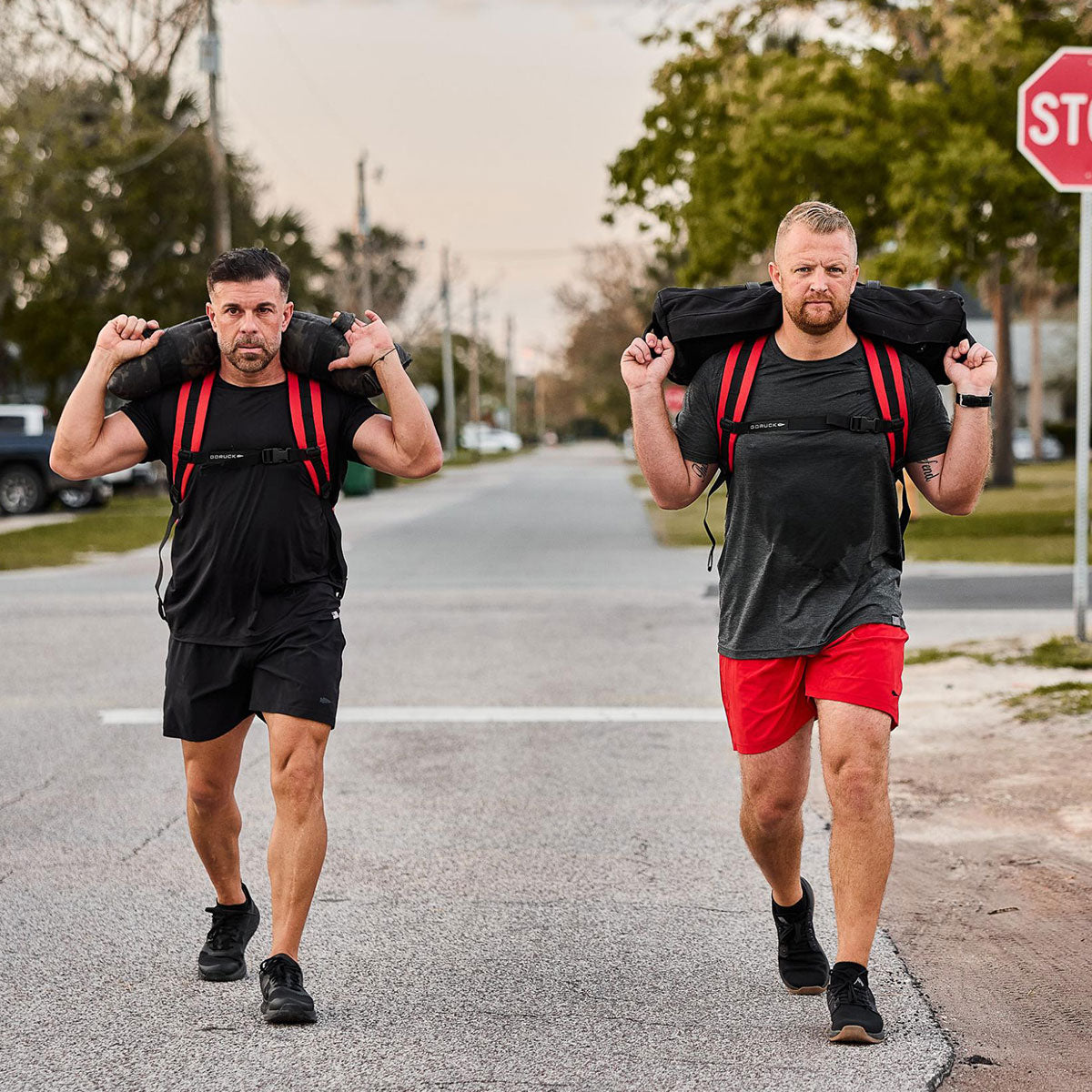 Two men carrying sandbags on their shoulders walk down a street wearing athletic gear.