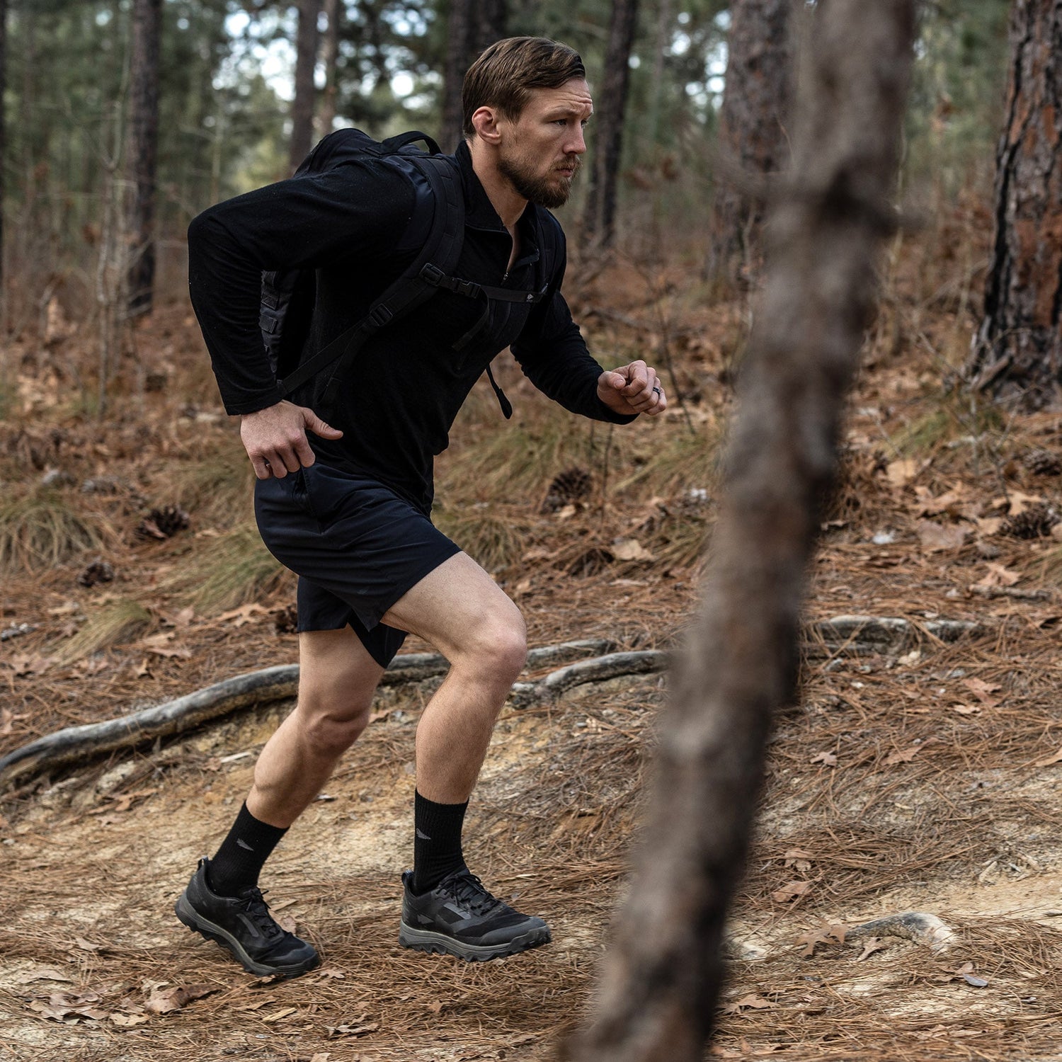 Man wearing black athletic gear and backpack hikes up a forest trail among trees and fallen pine needles.