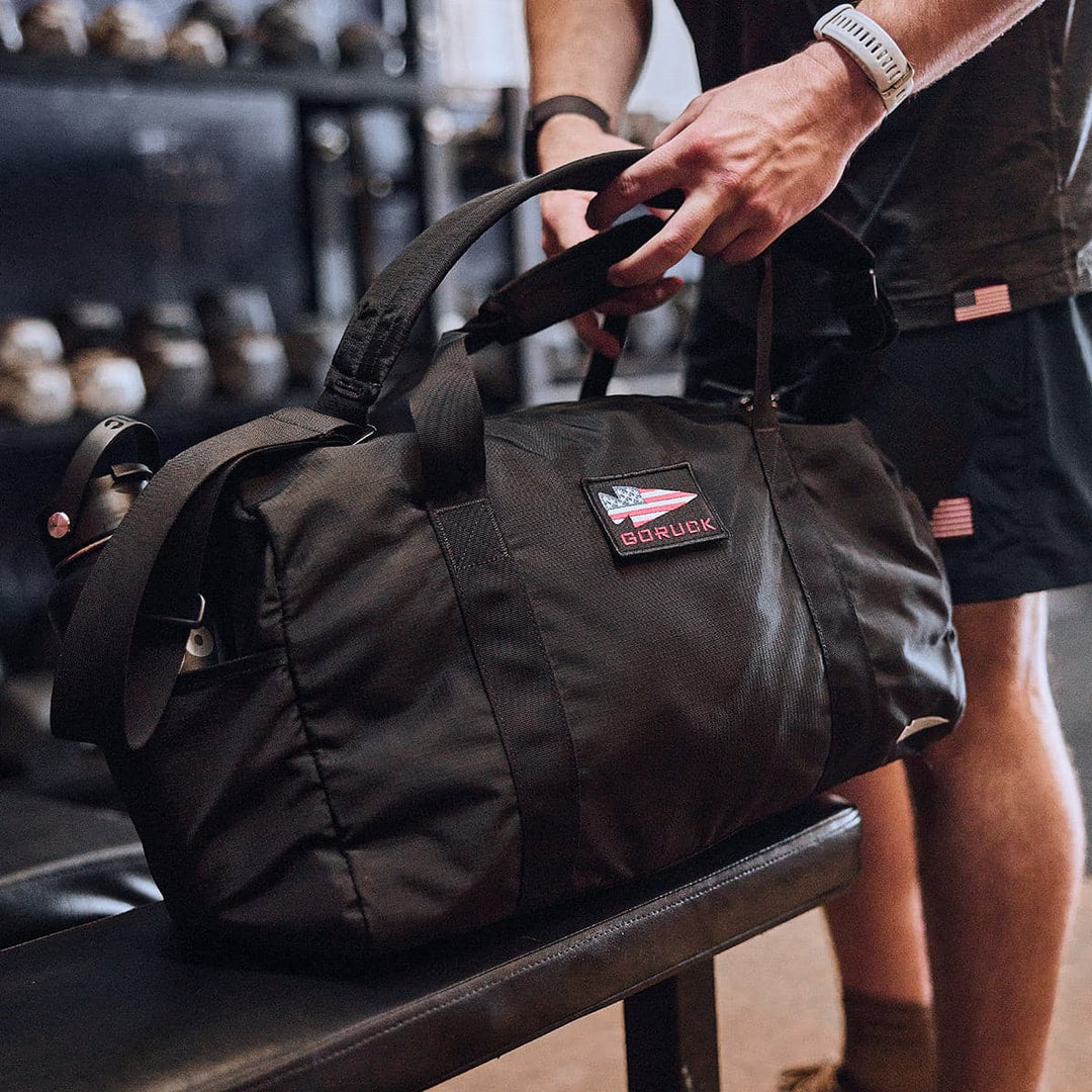A person wearing a smartwatch handles a large black GORUCK gym bag on a bench, showcasing the durable ripstop ROBIC® nylon material for lasting use. The bag also features a practical shoe compartment to maintain organization and separation of workout gear.
