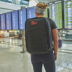 At the airport, a traveler with a carry-on compliant GR2 rucksack from GORUCK scans the flight information boards, confident in their Scars Lifetime Guarantee.