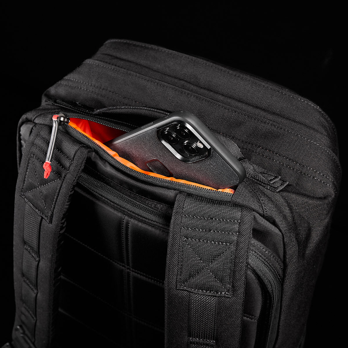 The GORUCK GR2, a black backpack featuring an orange-lined pocket that reveals part of a smartphone, takes cues from Special Forces medical packs for its rugged design. Set against a black background, this durable backpack comes with the reassurance of a Scars Lifetime Guarantee.