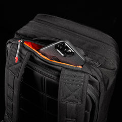 The GORUCK GR2, a black backpack featuring an orange-lined pocket that reveals part of a smartphone, takes cues from Special Forces medical packs for its rugged design. Set against a black background, this durable backpack comes with the reassurance of a Scars Lifetime Guarantee.