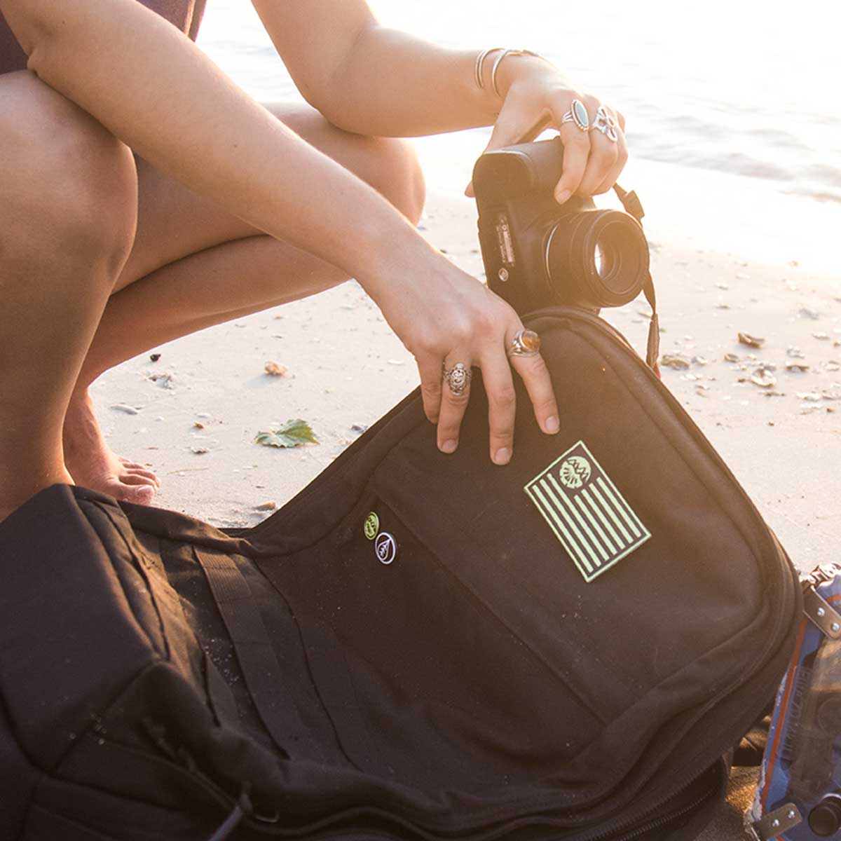 A person kneels on the beach, placing their camera into a black GORUCK backpack adorned with various patches. This GR2 pack, supported by the Scars Lifetime Guarantee, provides outstanding durability for every adventure.