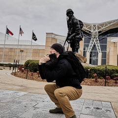 Wearing the Rucker - Long Range, a man in a beanie squats before a soldier statue and three flags, ready for distance rucking or upcoming ruck events.