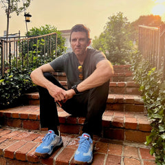 A man sits on brick steps outdoors at sunset, wearing a gray shirt, black pants, and the Mackall 45 Americana blue sneakers.