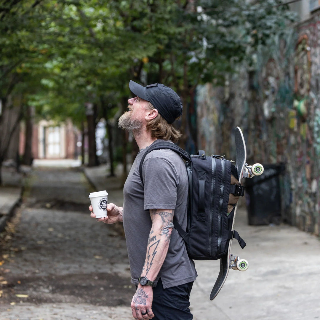 Man with beard wearing GORUCK backpack carrying skateboard and coffee in urban alleyway