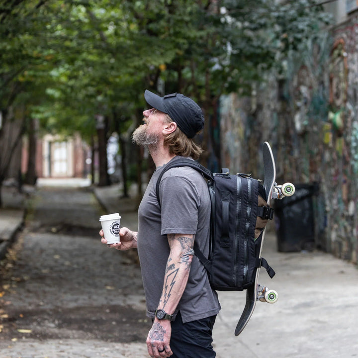 Man with beard wearing GORUCK backpack carrying skateboard and coffee in urban alleyway