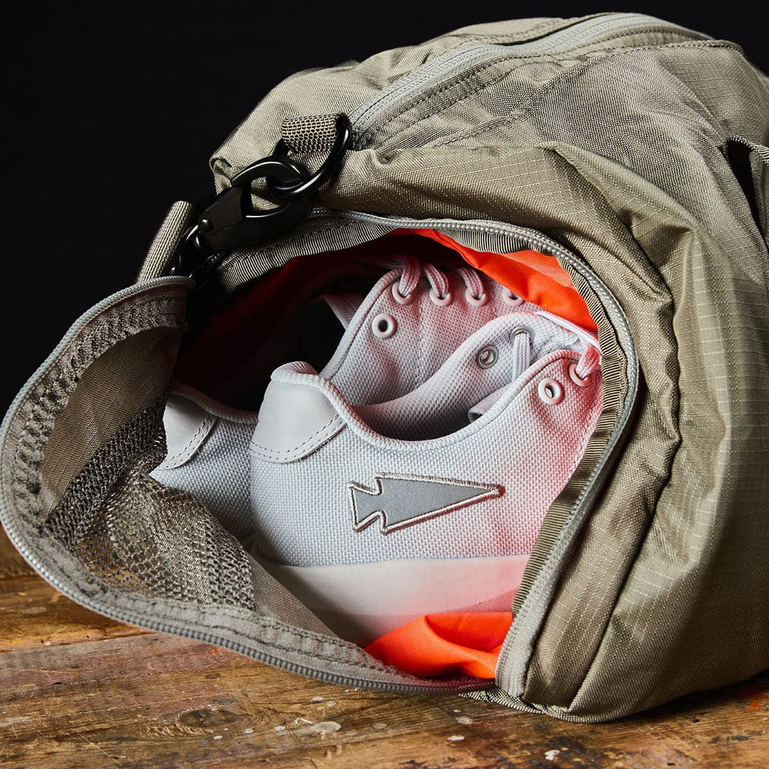 A pair of white sneakers with orange soles peeks out from the shoe compartment of a gray GORUCK Gym Bag - Ripstop ROBIC®, resting on a wooden surface.