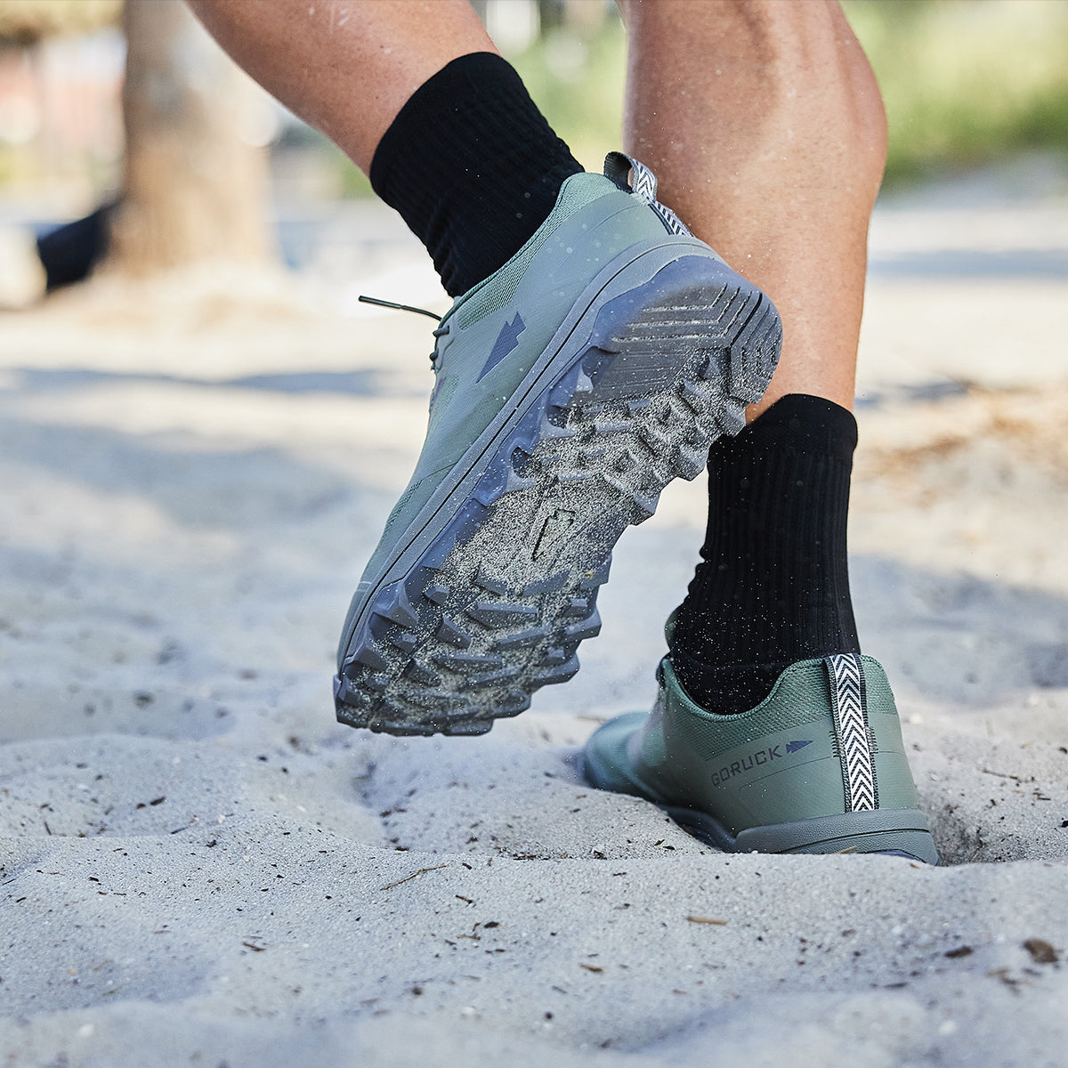 Close-up of a person running on sand, their feet clad in GORUCK's Mackall shoes in Earth and Forged Iron colors, paired with black socks, ideal for Special Forces training.