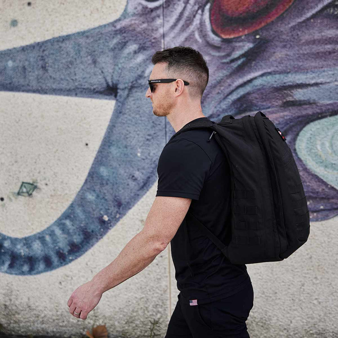 A man with a GORUCK GR2 backpack, renowned for its Scars Lifetime Guarantee, walks by a mural of a blue octopus.