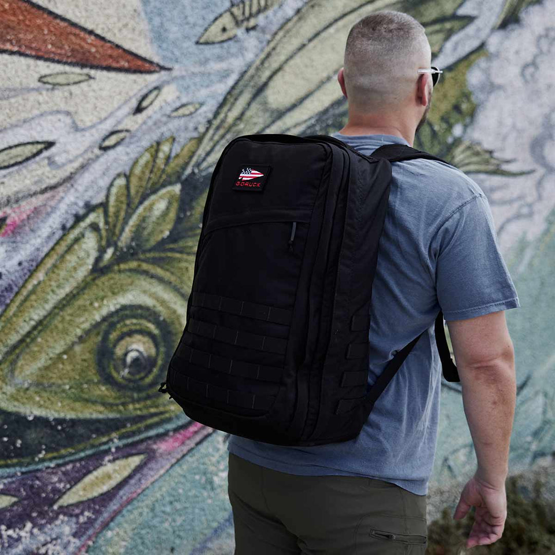 A person carrying a GORUCK GR2 backpack, possibly inspired by Special Forces medical packs, stands in front of a vibrant fish mural.