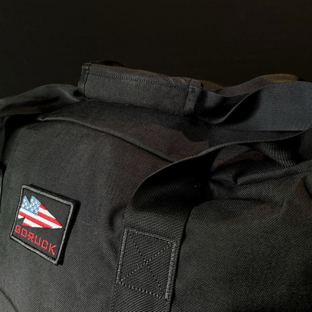 A dark-themed Kit Bag - 84L by GORUCK, featuring an American flag patch and "GORUCK" label, reminiscent of a deployment bag.