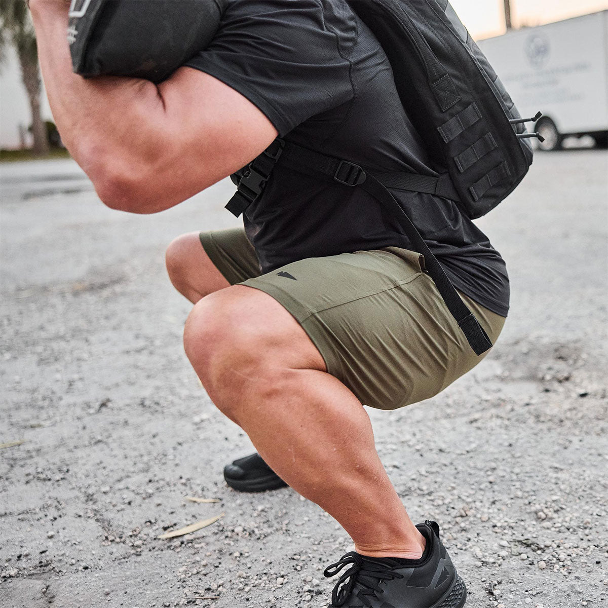 A person in workout gear squats outdoors while carrying a heavy sandbag on their shoulders.