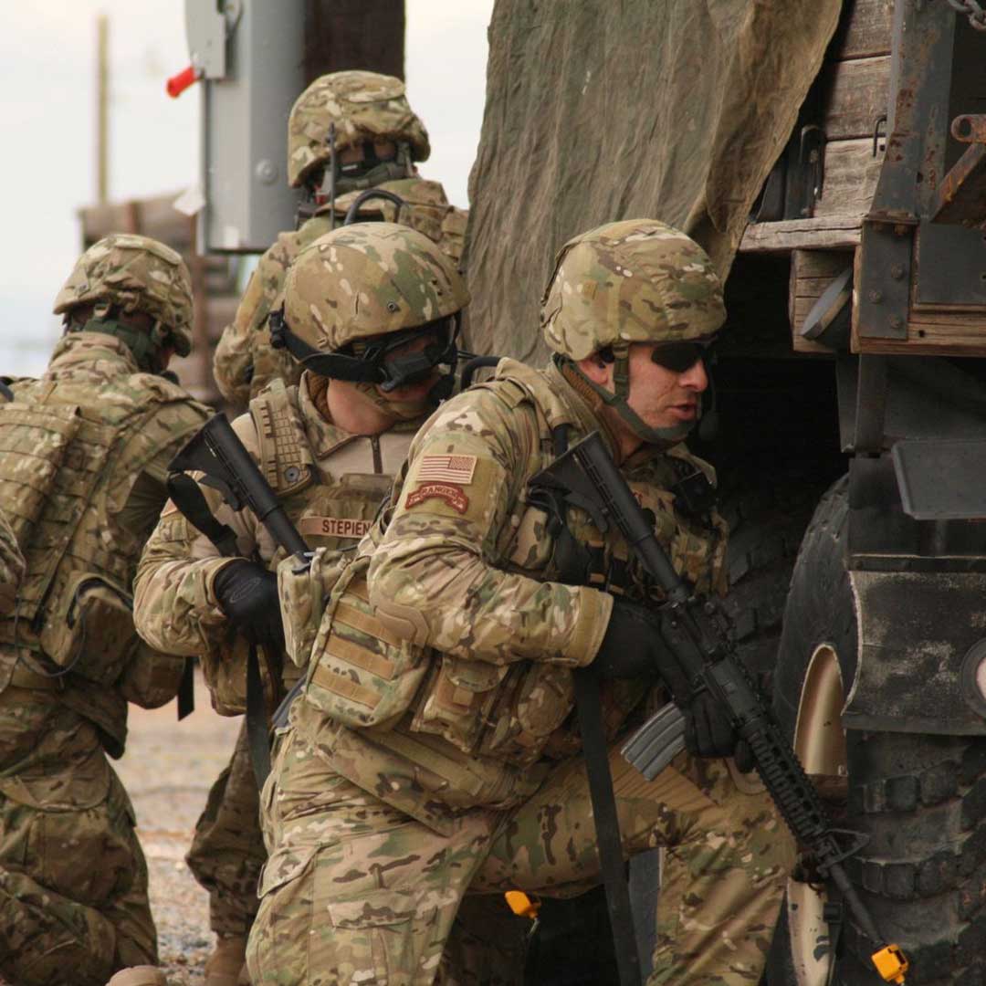 Four soldiers in camouflage gear and helmets take cover beside a military truck, holding rifles.