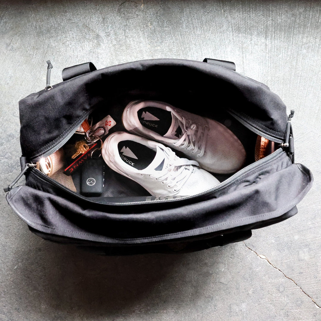 A GORUCK Kit Bag, outfitted with a shoulder strap and resembling a Special Forces Flight Bag, contains a pair of gray sneakers, a set of keys, a wallet, and a water bottle, all resting on a textured gray floor.