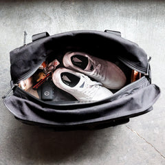 A GORUCK Kit Bag, outfitted with a shoulder strap and resembling a Special Forces Flight Bag, contains a pair of gray sneakers, a set of keys, a wallet, and a water bottle, all resting on a textured gray floor.