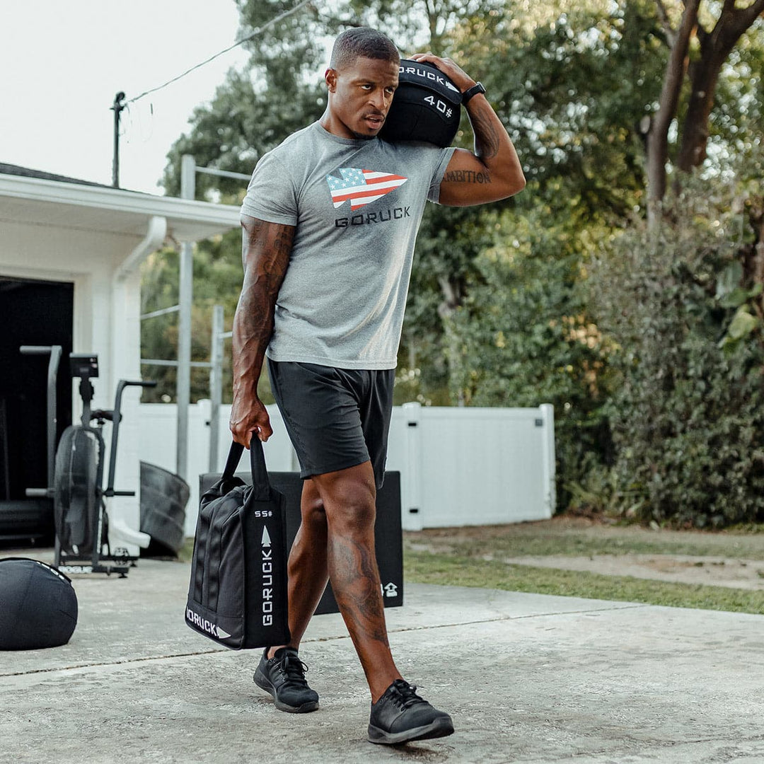 A person utilizes the Sand Jerry Can by GORUCK, made from Special Forces Grade materials, to exercise outdoors. They carry the weighted bags in both hands while wearing athletic attire near their home gym, concentrating on strengthening their core.