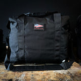 A black Kit Bag - 84L from GORUCK, featuring a design inspired by the Special Forces Flight Bag, is adorned with an American flag patch and rests on a wooden surface against a dark background.