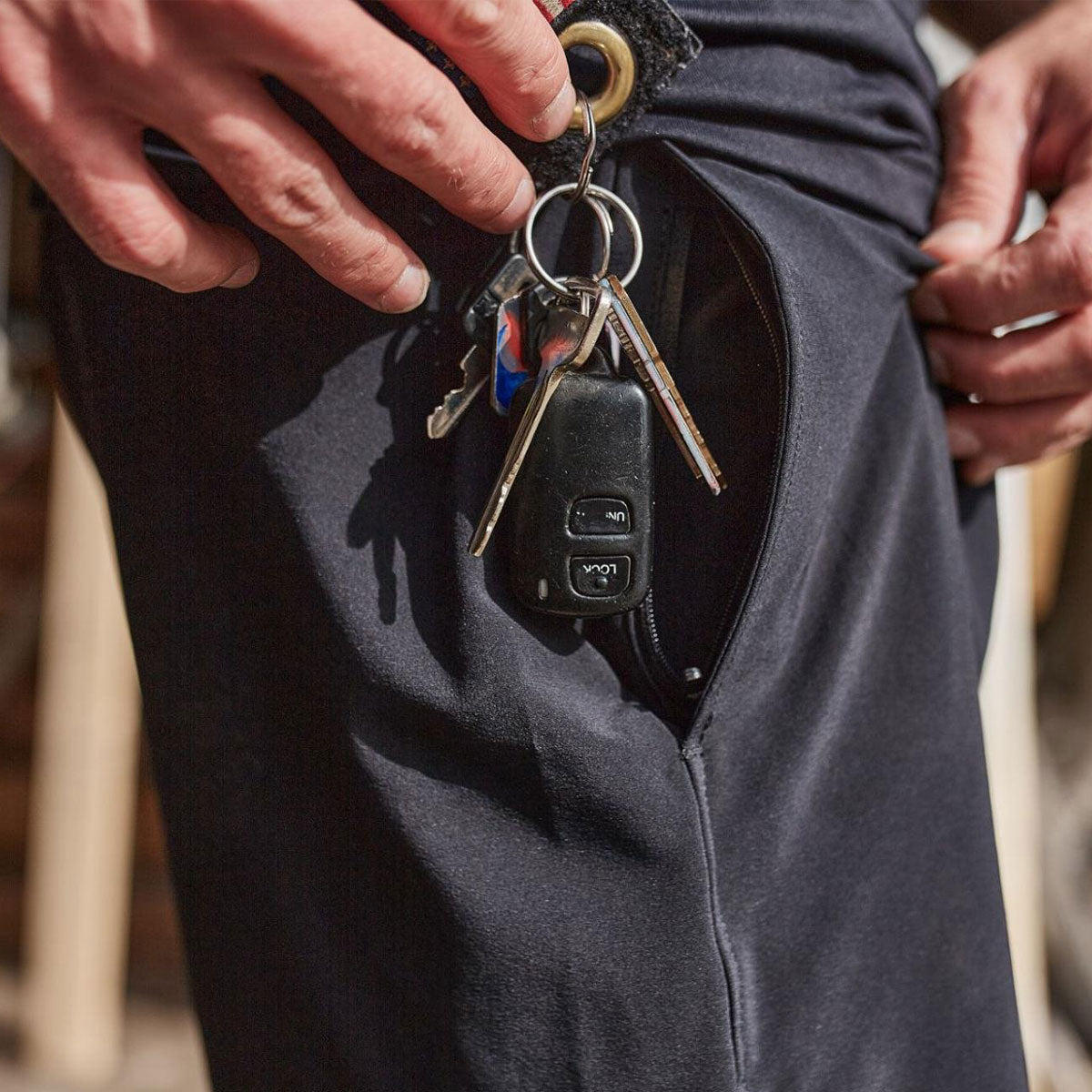 A hand puts a set of keys with a car fob into a zippered pocket on black pants.