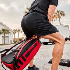Athlete lifting GORUCK red rucksack outdoors, wearing black shorts and shirt
