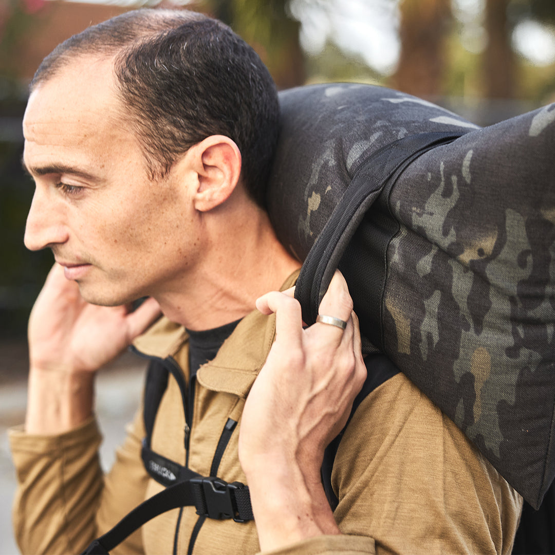 A man carries a large GORUCK Simple Training Sandbag with heavy-duty handles on his shoulder while wearing a brown jacket and backpack, exploring the outdoors.