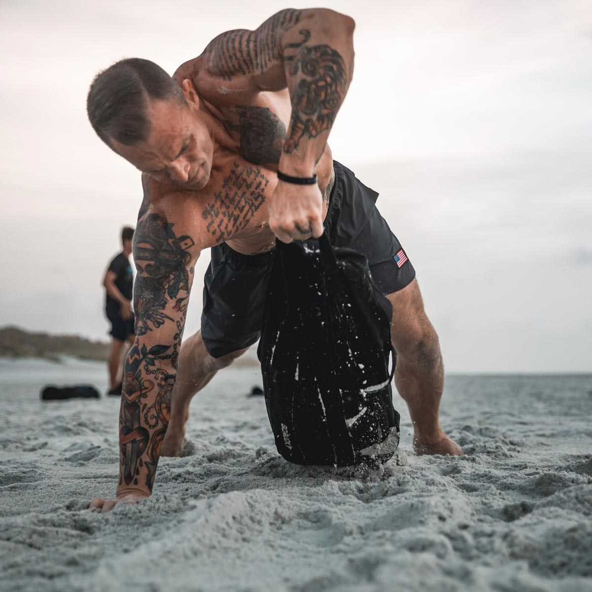 Tattooed man on the beach lifting a sandbag, with sand flying from the bag.