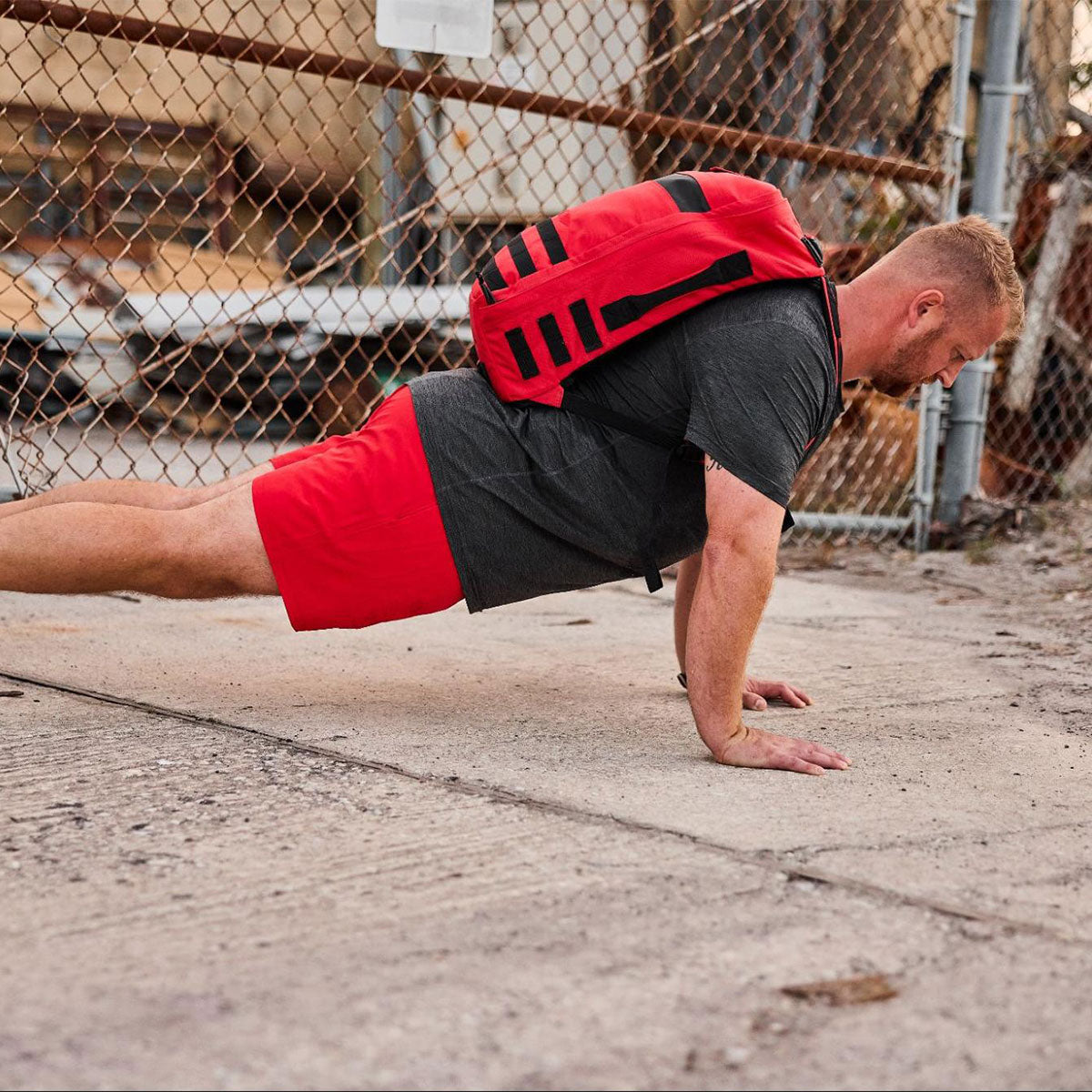 Man wearing a red backpack and red shorts doing a push-up on a concrete surface near a chain-link fence.