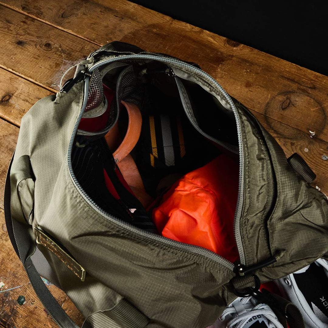 The open GORUCK Gym Bag - Ripstop ROBIC® rests on the wooden floor, showcasing its durable construction while filled with sports gear, including shoes neatly placed in the shoe compartment, a helmet, and an orange jacket.