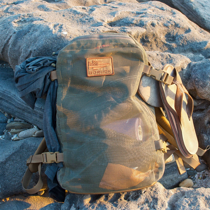 The Mesh Ruck backpack with a GORUCK patch, sandals, and clothing sits on sunlit rocks, drying quickly thanks to its breathable mesh design.