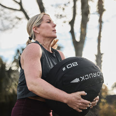 A woman outdoors, enhancing her core stability, lifts a large GORUCK Sand Medicine Ball during an invigorating workout.