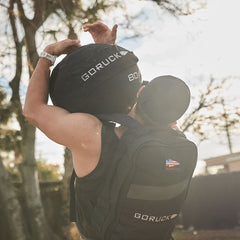 Dressed in workout attire, an individual lifts a GORUCK Sand Medicine Ball overhead outdoors, demonstrating their core stability while carrying a backpack adorned with a patch.