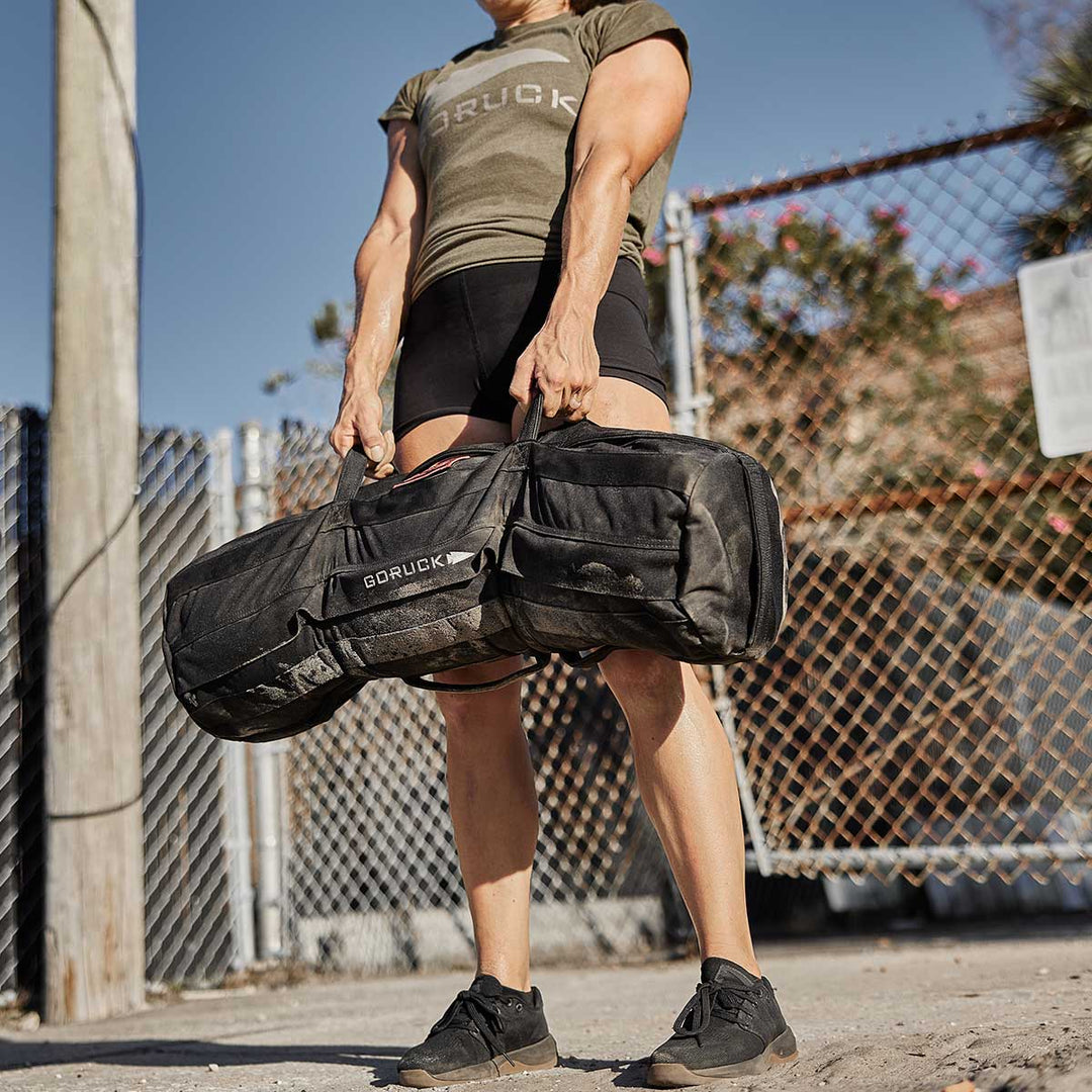 Wearing athletic gear, a person confidently lifts the GORUCK Sandbag Kit (w/ Filler Bag) with heavy-duty handles outdoors near a fence, pushing their limits.