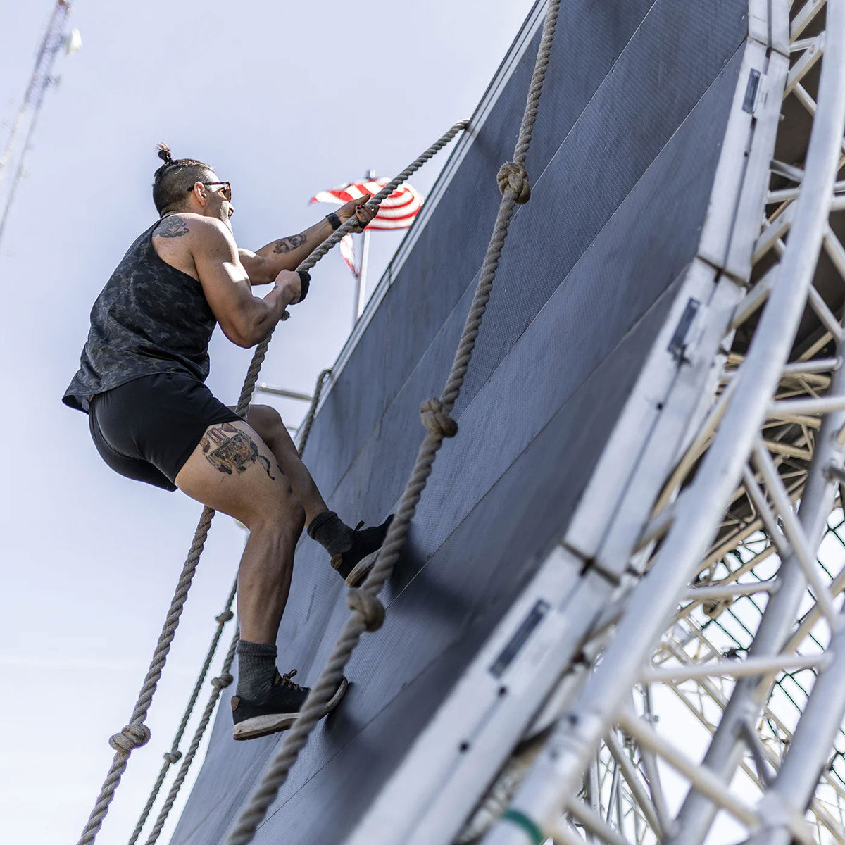 Man climbing a rope wall obstacle outdoors with a US flag in the background.