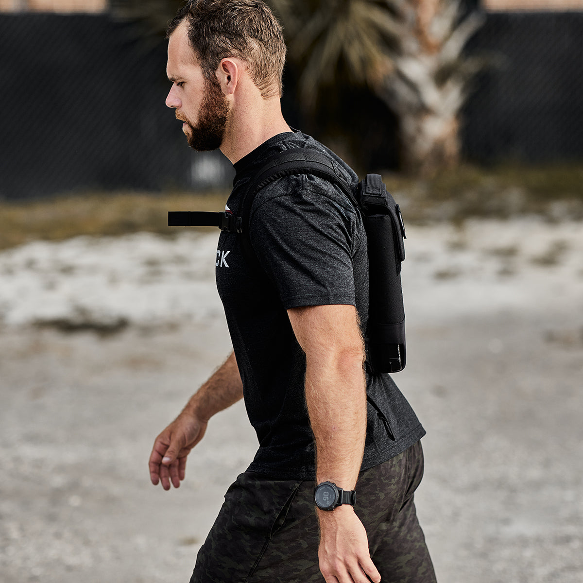 Man walking outdoors wearing a black t-shirt, camo shorts, and a hydration pack on his back.