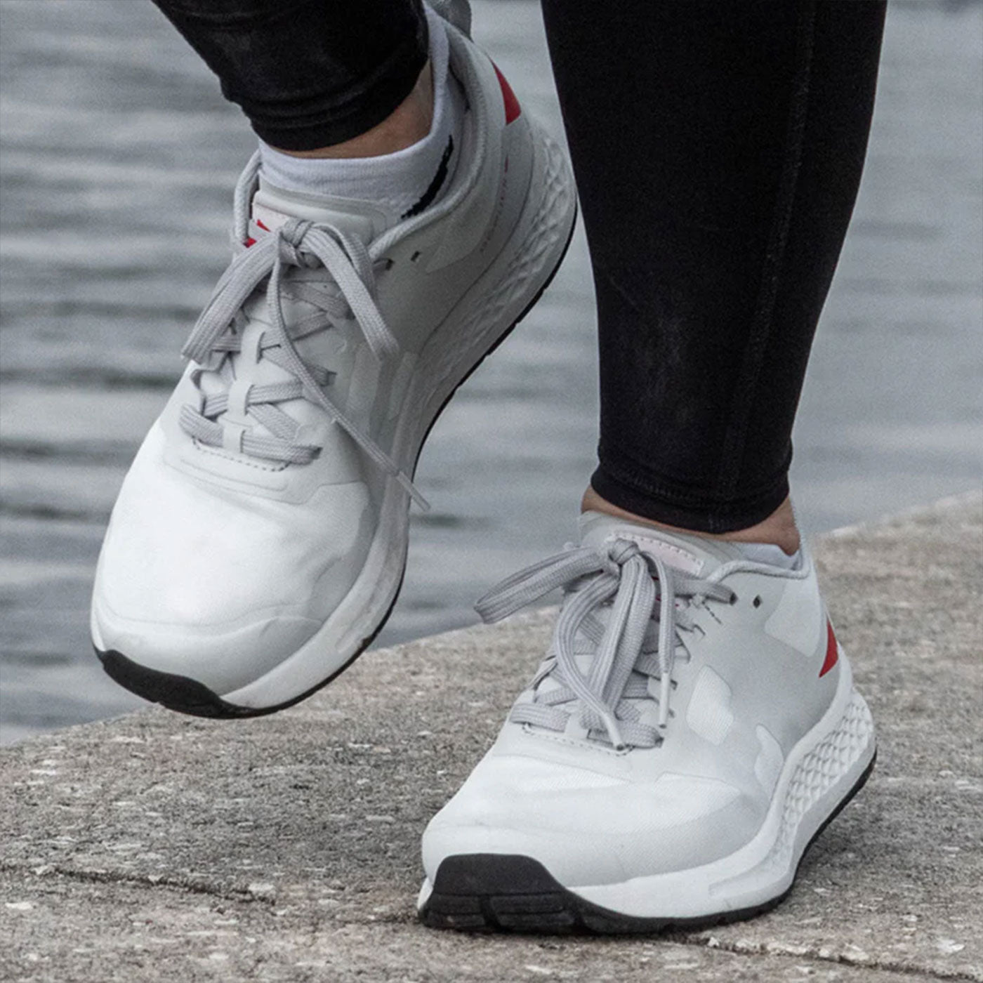 Person wearing white running shoes and black leggings walking on a concrete path by the water.