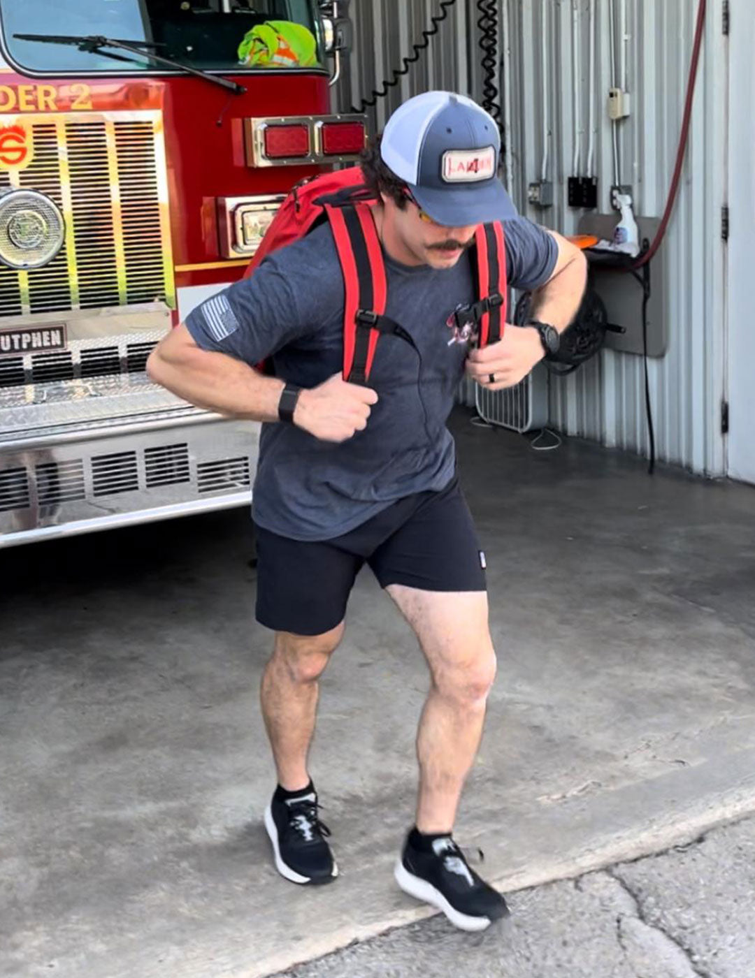 Man in athletic gear with a red backpack walks out of a fire station beside a parked fire truck.