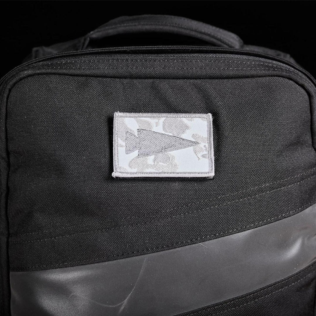 A black backpack with a white patch featuring a gray arrow design on the front.