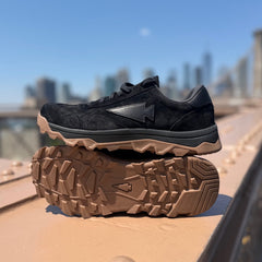 Mackall 45 Black + Gum sneakers with brown rugged soles displayed outdoors on a bridge with a city skyline in the background.